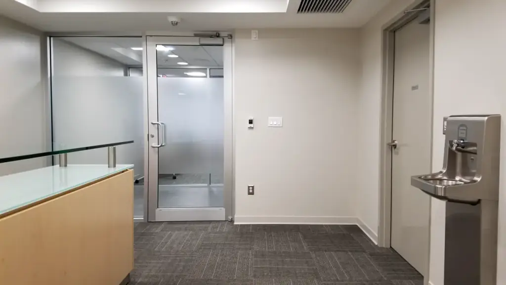 Office reception area with a glass-topped desk, glass entry door, wall plates, carpet tile, a restroom door and a water fountain